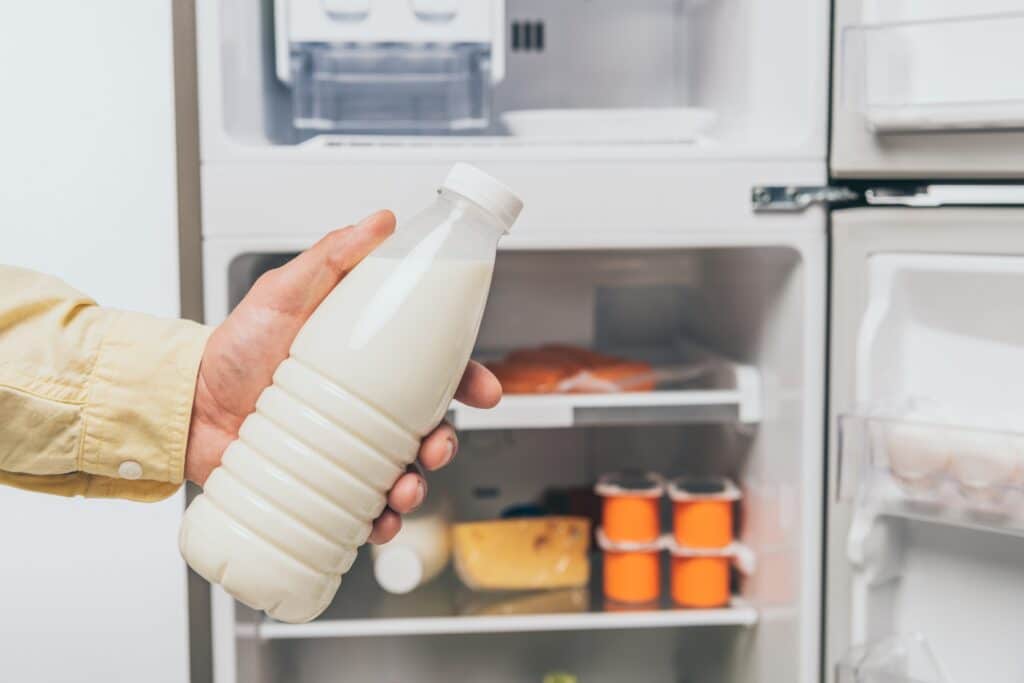 cropped view of man holding bottle of milk near open fridge with fresh food on shelves isolated on